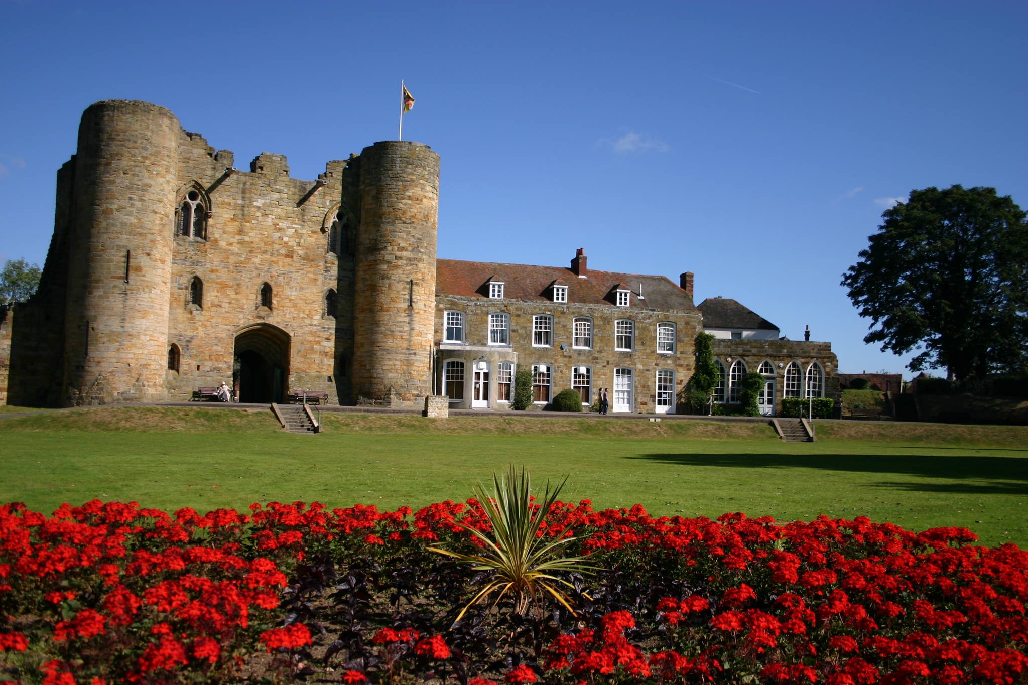 Tonbridge Castle Gatehouse and the mansion (now used as council offices) on a sunny day. In front or the mansion is a lawn and lots of flowers.